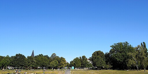 Volleyballfeld im Volkspark Friedrichshain  Volleyballfeld auf Sand mit Spielern im Vordergrund, umgeben von Rasenflächen, auf denen Leute liegen oder spazieren. Ein Weg führt durch den Park, Bäume und ein Kirchturm ragen im Hintergrund in den blauen Himmel.