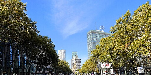 Blick über den Kurfürstendamm  Eine breite Allee mit Fußgängerbereich in der Mitte, gesäumt von Bäumen und modernen Gebäuden. Im Hintergrund das markante Hochhaus mit Mercedes-Stern.