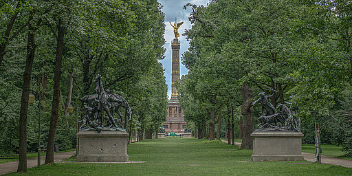 Ein breiter, von Bäumen gesäumter Weg im Berliner Tiergarten führt zur goldenen Siegessäule, flankiert von zwei großen Reiterskulpturen.