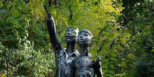  Zwei metallene Figuren, die beide mit Graffiti überzogen sind, stehen in einem dicht bewachsenen, grünen Park. Die Figuren schauen in unterschiedliche Richtungen und wirken wie abstrahierte Menschen mit verzierten Körpern. Die Umgebung ist stark bewachsen, mit überhängenden Ästen und Grünpflanzen, die das Bild dominieren. 