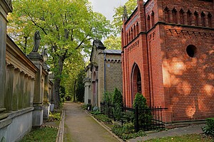 Ein Weg auf dem Alten St. Matthäus Kirchhof, flankiert von kunstvoll gestalteten Grabmälern. Links eine Reihe klassischer Grabstrukturen aus Sandstein, rechts eine rot geklinkerte Gruftkapelle mit gotischen Elementen. Der Pfad führt unter schattenspendenden grünen Bäumen entlang.
