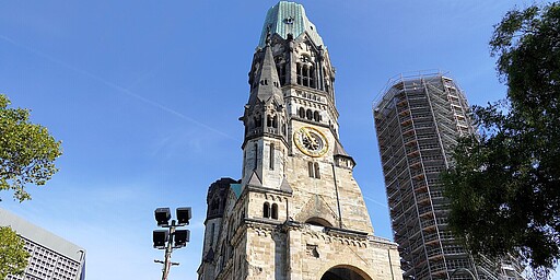 Nahaufnahme der Kaiser-Wilhelm-Gedächtniskirche  Eine Nahaufnahme der Kaiser-Wilhelm-Gedächtniskirche, die den beschädigten Turm in all seinen Details zeigt. Der Turm hat eine imposante Uhr, die gut sichtbar ist, und das steinerne Mauerwerk ist teilweise beschädigt und zeigt Spuren des Krieges. Rechts daneben ist ein moderner, eingerüsteter Turm zu sehen. Der klare blaue Himmel verleiht dem Bild eine friedliche Atmosphäre, trotz der Mahnung, die das Gebäude darstellt.