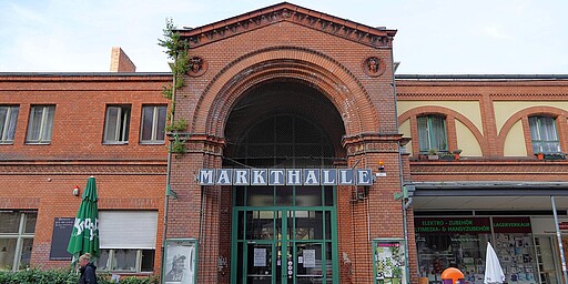Fassade der Arminiusmarkthalle Die historische Arminiusmarkthalle mit ihrer roten Backsteinfassade und dem großen Bogeneingang. Über dem Eingang steht in großen Buchstaben „Markthalle“. Links ist ein grüner Sonnenschirm und eine kleine Begrünung zu sehen.