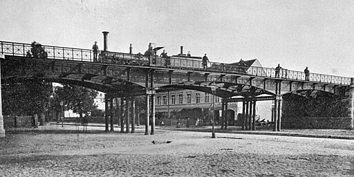Brücke Müllerstraße Ringbahnbrücke über die Müllerstraße am Bahnhof Wedding: Auf der Brücke steht eine 2B-Lok, die zur aus den USA bezogenen Erstausstattung der Niederschlesisch-Märkischen-Eisenbahn gehört. Ohne Witterungsschutz für den Lokführer und mit dem riesigen Stehkessel ist sie vermutlich in den 1850er-Jahren entstanden und ähnelt (bis auf die Achsfolge) der Lok „Beuth“ von Borsig (1843 gebaut).