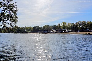 Ein weiter Blick über den Plötzensee in Berlin. Das glitzernde Wasser spiegelt die Sonne wider, während am Ufer Bäume und ein kleiner Strand zu sehen sind. Einige Gebäude und weiße Zelte ergänzen die Szenerie.