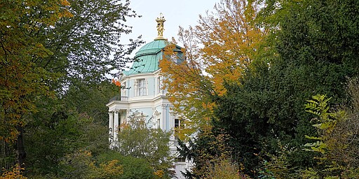 Verstecktes Belvedere im herbstlichen Grün  Das Belvedere im Schlossgarten Charlottenburg ist von herbstlich gefärbtem Laub und hohen Bäumen umrahmt. Die hellblaue Fassade des Gebäudes schimmert durch das dichte Grün. Auf dem Dach ist eine goldene Figur sichtbar, die von einem kupfernen Kuppeldach gekrönt wird. Ein schmaler Weg führt durch das Gebüsch in Richtung des Gebäudes.