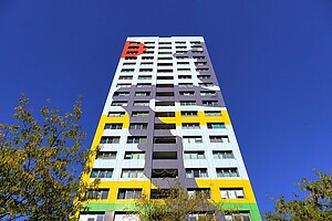 Ein Hochhaus mit einer künstlerischen Fassadengestaltung, bestehend aus bunten, geometrischen Formen in Gelb, Lila, Grün und Rot. Das Design wirkt wie ein riesiges abstraktes Gemälde. Der klare blaue Himmel hebt die Farben noch mehr hervor. Baumkronen am unteren Bildrand ergänzen das Bild.