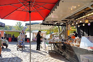 Eine entspannte Szene auf dem Flohmarkt am Mauerpark, unter einem großen roten Sonnenschirm. Ein Mann schiebt einen Kinderwagen, während eine Frau an einem Stand für Süßwaren und Backwaren stöbert.