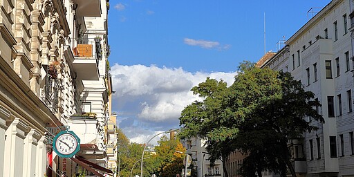 Akazienkiez bei Sonnenschein Eine belebte Straße im Akazienkiez, mit alten Berliner Mietshäusern, deren Balkone mit Pflanzen geschmückt sind. Im Vordergrund ist ein Antiquitätengeschäft zu sehen, an dem Kleidung draußen hängt. Der Himmel ist klar, und die Bäume an der Straße sind voll belaubt. Menschen spazieren die Straße entlang.