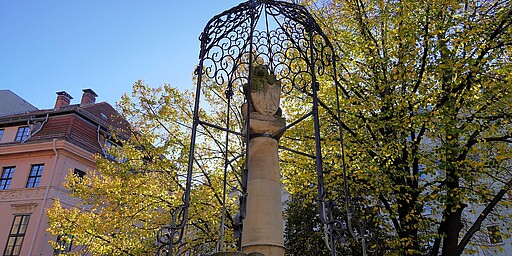 Ein dekorativer Brunnen mit einer verzierten Metallkuppel steht vor einem herbstlichen Baum im Nikolaiviertel. Die Blätter schimmern in sattem Gelb, während das Sonnenlicht durch das filigrane Metallgeflecht des Brunnens scheint. Im Hintergrund ist ein Altbau in zarten Rosatönen zu sehen.