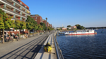 Ein sonniger Tag an der Promenade der Rummelsburger Bucht in Berlin. Rechts verläuft ein Gehweg entlang des Wassers mit Bäumen, die Schatten spenden. Links befinden sich moderne Backsteingebäude mit Balkonen, dahinter liegt das Wasser, auf dem ein Ausflugsboot fährt. Menschen spazieren entlang des Ufers und genießen den Blick auf die Bucht.