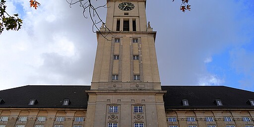 Rathaus Schöneberg – Stolz mit Turm Nahaufnahme des Turms des Rathauses Schöneberg in Berlin, der in den Himmel ragt. Der Turm hat eine große Uhr mit römischen Ziffern und ist umgeben von einem neoklassizistischen Gebäude. Über dem Turm hängen herbstliche Äste mit orangefarbenen Blättern. Der Himmel ist bewölkt, lässt aber auch etwas blau durchblicken.