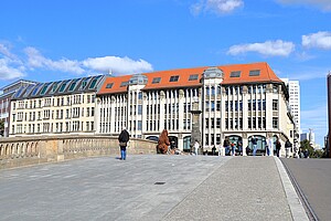 Touristen und Berliner schlendern über eine Brücke auf der Museumsinsel, flankiert von imposanten Gebäuden, unter strahlend blauem Himmel.