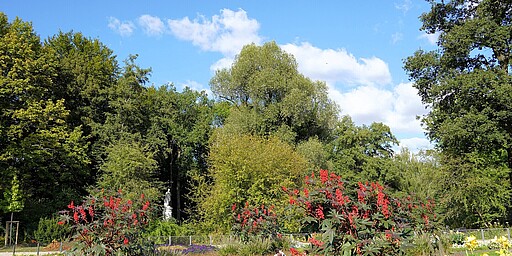 Bunter Garten auf der Luiseninsel Ein gepflegter Garten in Berlin, Luiseninsel im Großen Tiergarten. Bunte Blumenbeete mit roten, lila und gelben Blumen umgeben von hohen grünen Bäumen. Im Hintergrund eine weiße Statue. Eine Person sitzt im Gras, genießt die Sonne.