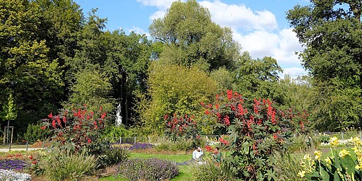  Ein gepflegter Garten in Berlin, Luiseninsel im Großen Tiergarten. Bunte Blumenbeete mit roten, lila und gelben Blumen umgeben von hohen grünen Bäumen. Im Hintergrund eine weiße Statue. Eine Person sitzt im Gras, genießt die Sonne. 