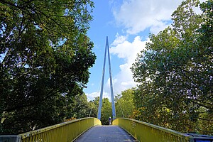 Eine moderne gelbe Fußgängerbrücke, umrahmt von dichten grünen Bäumen. Im Hintergrund ragt eine metallene, dreieckige Struktur in den blauen Himmel. Ein Mensch geht über die Brücke.