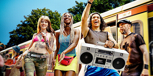 Bund und wild Feiernde Personen in bunter Kleidung mit einer Musikanlage vor der S-Bahn.