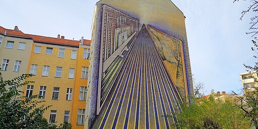 Abstrakte 3D-Wandmalerei in Berlin  Die Hausfassade zeigt ein abstraktes Wandbild mit geometrischen Mustern und Linien, die in die Ferne führen. Die Farben und die Perspektive schaffen einen lebendigen, dreidimensionalen Eindruck auf der sonst flachen Fläche.