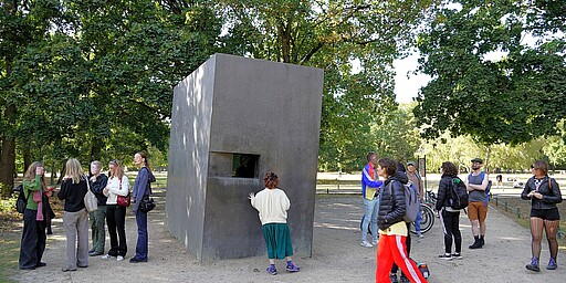 Denkmal der Stille im Tiergarten Eine Gruppe von Menschen steht um das Denkmal für die im Nationalsozialismus verfolgten Homosexuellen im Berliner Tiergarten. Das Denkmal besteht aus einem schlichten, kantigen Betonkubus mit einer kleinen, rechteckigen Öffnung, in die eine Person hineinblickt. Die Gruppe besteht aus Männern und Frauen, einige stehen im Gespräch, andere blicken nachdenklich auf das Denkmal. Umgeben ist die Szene von hohen, grünen Bäumen, die das Denkmal in eine ruhige und friedliche Atmosphäre einbetten.