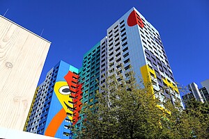 Ein Hochhaus mit auffälliger, farbenfroher Fassadengestaltung in leuchtendem Blau, Gelb, Rot und weiteren Farben. Die Gestaltung zeigt abstrakte Muster und Formen. Im Vordergrund sind Baumkronen mit grünem Laub zu sehen, die das Gebäude einrahmen. Der Himmel ist klar und tiefblau.