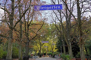 Eine Allee von Bäumen mit leuchtend buntem Herbstlaub, darunter ein Biergarten mit Girlanden und Bänken. Menschen spazieren entspannt im Hintergrund. Eine gemütliche Atmosphäre liegt in der Luft.