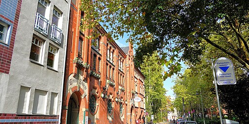 Straßenszene vor dem Städtischen Volksbad Charlottenburg  Eine ruhige Straße in Berlin-Charlottenburg. Links stehen Wohnhäuser mit roten und grauen Fassaden, die von Bäumen gesäumt sind. Im Zentrum ist die prächtige Backsteinfassade des ehemaligen Volksbads Charlottenburg mit verzierten Fenstern und einem großen Torbogen zu sehen. Rechts ist ein Parkscheinautomat und ein paar parkende Fahrräder und Autos. Die Szene wird von Herbstbäumen eingerahmt.