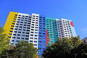 Ein großes, modernes Hochhaus mit vielen Balkonen, bemalt in bunten Farben. Links gelb, in der Mitte blau und grün, rechts weiß mit einem roten Kreis. Vor dem Gebäude stehen einige Bäume, und der Himmel ist strahlend blau.