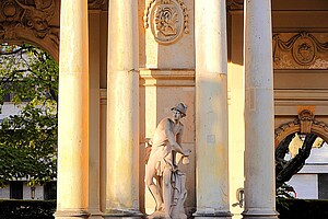 Eine steinerne Statue eines Mannes in antiker Gewandung, flankiert von hohen, hellen Marmorsäulen. Die Szene wird von warmem Sonnenlicht erleuchtet, wodurch die Details der Säulen und der Statue hervorgehoben werden. Im Hintergrund schimmert grünliches Laub durch.