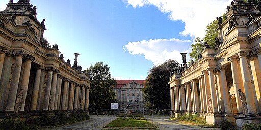  Die Königskolonaden im Heinrich-von-Kleist-Park, zwei symmetrische Säulengänge mit kunstvollen Statuen auf den Dächern. In der Mitte des Bildes ein historisches Gebäude und ein blauer Himmel mit vereinzelten Wolken. 