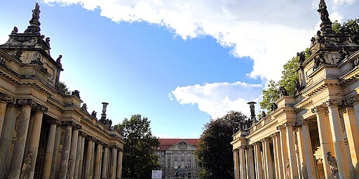 Königskolonaden im Heinrich-von-Kleist-Park Die Königskolonaden im Heinrich-von-Kleist-Park, zwei symmetrische Säulengänge mit kunstvollen Statuen auf den Dächern. In der Mitte des Bildes ein historisches Gebäude und ein blauer Himmel mit vereinzelten Wolken.