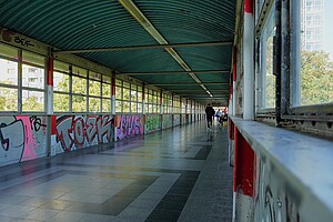 Ein langgezogener, überdachter Fußgängerkorridor über einer Straße ist zu sehen, die Wände sind mit bunten Graffiti verziert. Der Boden ist mit quadratischen Kacheln belegt und einige Menschen gehen durch den Gang.