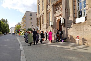 Eine Gruppe von festlich gekleideten Menschen steht vor dem Eingang eines historischen Gebäudes. Der Eingang ist mit großen Säulen und Skulpturen geschmückt. Der Himmel ist blau, und das Licht des Nachmittags betont die Farben der Szene.