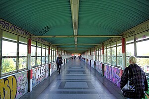 Ein langer, überdachter Fußgängertunnel mit türkisfarbener Decke und großen Fenstern an den Seiten. Die Wände sind mit buntem Graffiti verziert. Mehrere Personen laufen durch den Tunnel, eine ältere Frau im Mantel ist von hinten zu sehen, während ein Mann in dunkler Kleidung auf den Betrachter zuläuft. Draußen sind Bäume und Gebäude zu erkennen.