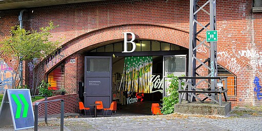 Eingang zum Buchstabenmuseum  Ein breiter Bogen aus rotem Backstein führt in das Buchstabenmuseum, das unter einer Eisenbahnbrücke liegt. Vor dem Eingang stehen zwei orangefarbene Stühle und ein Tisch, und an der Wand hängt ein leuchtend grünes Schild.