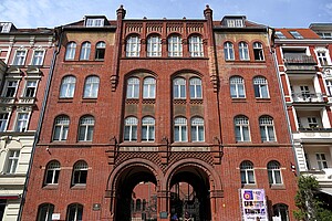 Die Fassade eines roten Backsteingebäudes, die Synagoge in der Rykestraße in Berlin. Das Gebäude hat mehrere hohe Fenster und zwei große, symmetrische Torbögen, die zum Innenhof führen. Rechts und links sind Wohnhäuser zu sehen, deren Fassaden aufwendig verziert sind. Ein Plakat für eine Veranstaltung ist rechts unten am Gebäude angebracht.