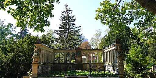 Alter Friedhof mit Denkmälern Ein alter Friedhof mit einem imposanten Denkmal im Vordergrund, umgeben von hohen Bäumen. Das Denkmal besteht aus einer steinernen Struktur mit Säulen und Statuen, die eine friedliche Atmosphäre inmitten der Natur ausstrahlt.