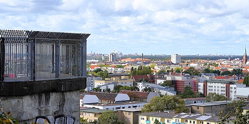 Aussicht vom Flakturm im Humboldthain Blick von einem Flakturm auf die Stadt Berlin. Ein Geländer mit Metallstäben am linken Bildrand, im Hintergrund erstreckt sich die Stadt mit Häusern, roten Dächern und einem Kirchturm, bei bewölktem Himmel.