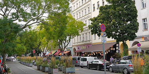 Die Bergmannstraße im Grünen Blick auf die Bergmannstraße, gesäumt von Bäumen, die Schatten auf die Straße werfen. Entlang der Gehwege stehen Autos geparkt, während Fußgänger und Fahrradfahrer die ruhige Atmosphäre genießen. Die Häuser auf der rechten Seite haben helle Fassaden und wirken gepflegt, teilweise mit Cafés und kleinen Läden.