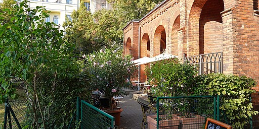 Café Strauss am Alten St.-Matthäus-Kirchhof Eine einladende Terrasse des Café Strauss, umgeben von blühenden Sträuchern und Bäumen. Die roten Backsteinbögen des Gebäudes bilden einen warmen Kontrast zur grünen Umgebung.