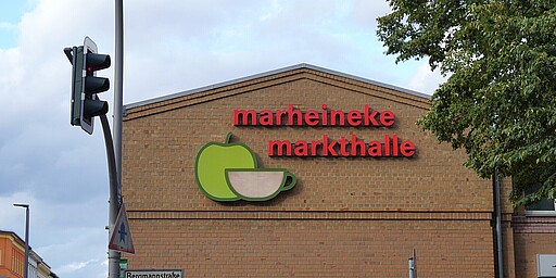 Detailansicht der Marheineke Markthalle Nahaufnahme des Giebels der Marheineke Markthalle mit der Aufschrift „marheineke markthalle“ in roten Buchstaben. Ein grünes Logo in Form eines Apfels mit einer Tasse ist neben dem Schriftzug angebracht. Vor der Halle stehen eine Ampel und Straßenschilder, darunter das Schild der „Bergmannstraße“.