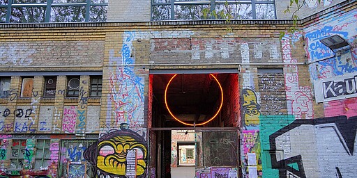 Eingang von Panke Culture Der Eingang von Panke Culture, einem Kulturzentrum in Berlin. Die Fassade ist mit bunten Graffitis verziert, darunter ein grinsendes gelbes Gesicht und andere künstlerische Elemente. Im Torbogen leuchtet ein roter Neonkreis.