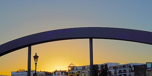 Sonnenuntergang über der Stadt  Eine Brücke im Vordergrund bildet einen dunklen Bogen über dem Bild, während im Hintergrund die untergehende Sonne den Himmel in warme Farben taucht. Im Hintergrund sind Wohngebäude und ein nostalgischer Straßenlaternenpfahl zu sehen.