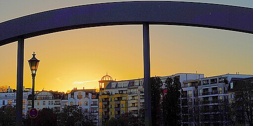  Eine Brücke im Vordergrund bildet einen dunklen Bogen über dem Bild, während im Hintergrund die untergehende Sonne den Himmel in warme Farben taucht. Im Hintergrund sind Wohngebäude und ein nostalgischer Straßenlaternenpfahl zu sehen. 