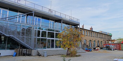 Blick auf das Zentrum für Kunst und Urbanistik Eine moderne Glasfassade des Zentrums für Kunst und Urbanistik (ZKU) in Moabit, flankiert von einem Baum mit herbstlich gefärbtem Laub. Im Hintergrund ein altes Industriegebäude aus gelbem Backstein, das den Charme des Ortes unterstreicht.