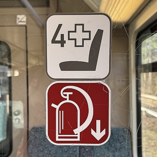 Auf dem Windfang im Eingangsbereich weist ein Feuerlöscher-Piktogramm (altes Design) den Weg zum in Bodennähe hinter einer Klappe verstauten Feuerlöscher. Das Piktogramm darüber bedeutet: 4 Sitzplätze für mobilitätseingeschränkte Fahrgäste