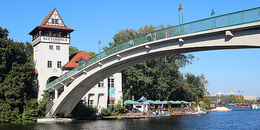 
    
        Ein Foto der Insel der Jugend in Berlin, aufgenommen an einem sonnigen Tag. Die Abteibrücke erstreckt sich über das Wasser und verbindet die Insel mit dem Ufer. Links im Bild ist das Kulturhaus der Insel zu sehen, ein großes weißes Gebäude mit einem steilen, roten Dach. Die Brücke ist leicht gewölbt und trägt Lampen. Darunter befindet sich ein Café mit Sonnenschirmen und Menschen am Wasser.
    
