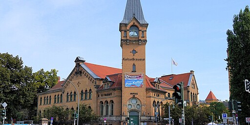 Die Kulturbrauerei in voller Pracht  Das Bild zeigt die Kulturbrauerei in Berlin, ein großes Gebäude im Backstein-Stil mit einem hohen, spitzen Turm. Vor dem Gebäude sind Straßenlaternen und einige Passanten zu sehen. Die Architektur ist typisch für die industrielle Bauweise des 19. Jahrhunderts.