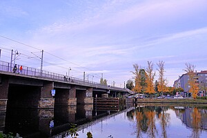 Eine Brücke über einen ruhigen Fluss mit goldgelben Laubbäumen im Hintergrund. Der Himmel ist in zarten Pastelltönen gefärbt.