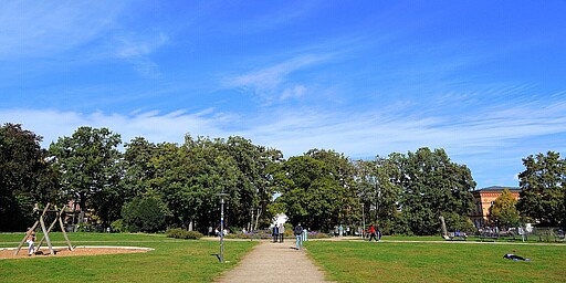  Ein breiter Kiesweg führt durch den Forckenbeckplatz. Links sieht man eine Holzschaukel auf einem Kinderspielplatz, und rechts erstrecken sich grüne Wiesen. Mehrere Menschen sind im Park zu sehen, einige stehen und unterhalten sich, andere spazieren. Am Horizont erstreckt sich ein blauer Himmel mit leichten, weißen Wolkenstreifen. 