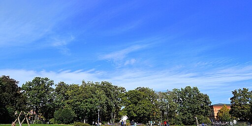 Spazierweg im Forckenbeckplatz bei strahlendem Himmel Ein breiter Kiesweg führt durch den Forckenbeckplatz. Links sieht man eine Holzschaukel auf einem Kinderspielplatz, und rechts erstrecken sich grüne Wiesen. Mehrere Menschen sind im Park zu sehen, einige stehen und unterhalten sich, andere spazieren. Am Horizont erstreckt sich ein blauer Himmel mit leichten, weißen Wolkenstreifen.
